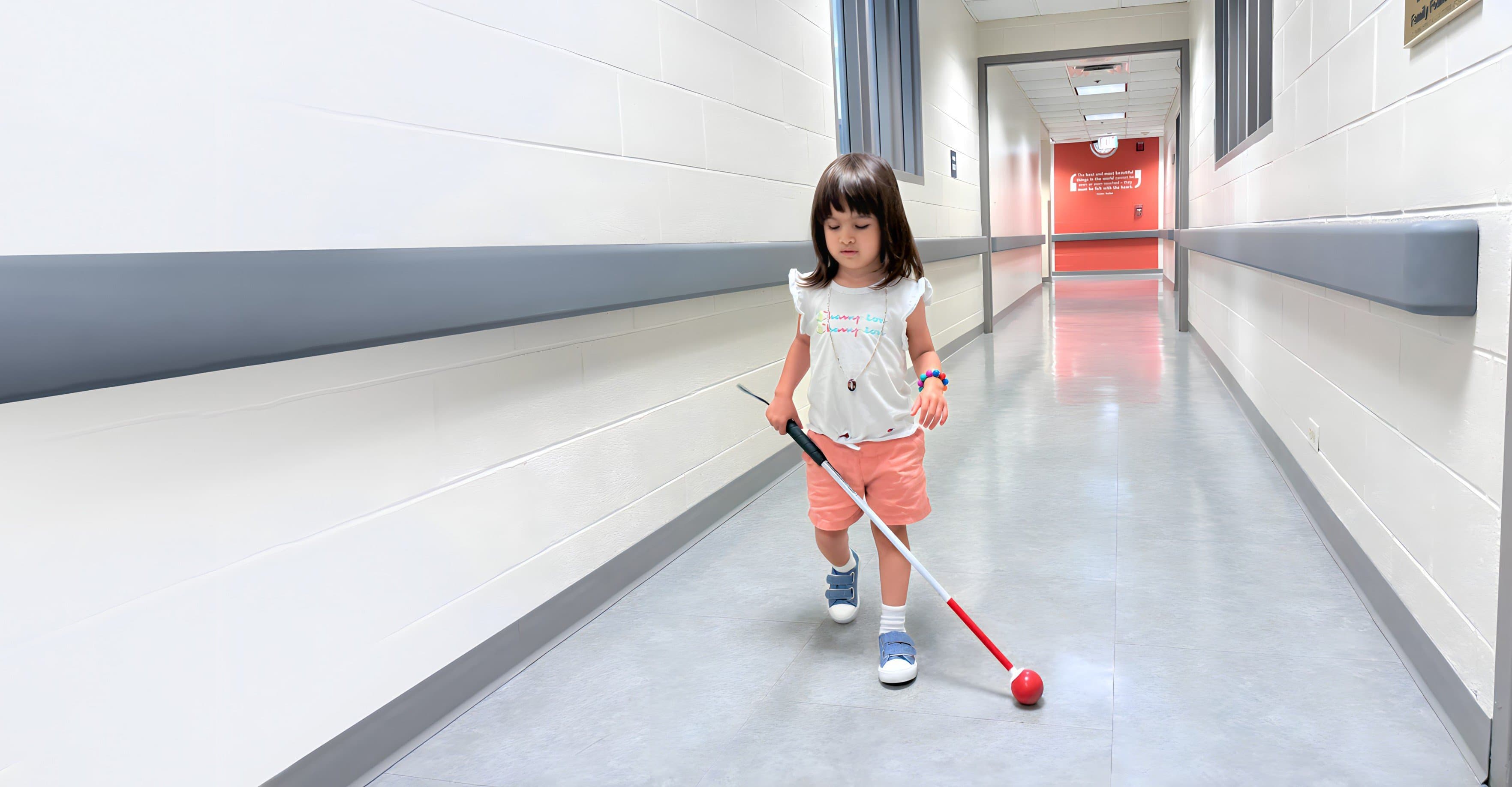 A little girl using a tiny white cane walks down a hallway, navigating by herself.