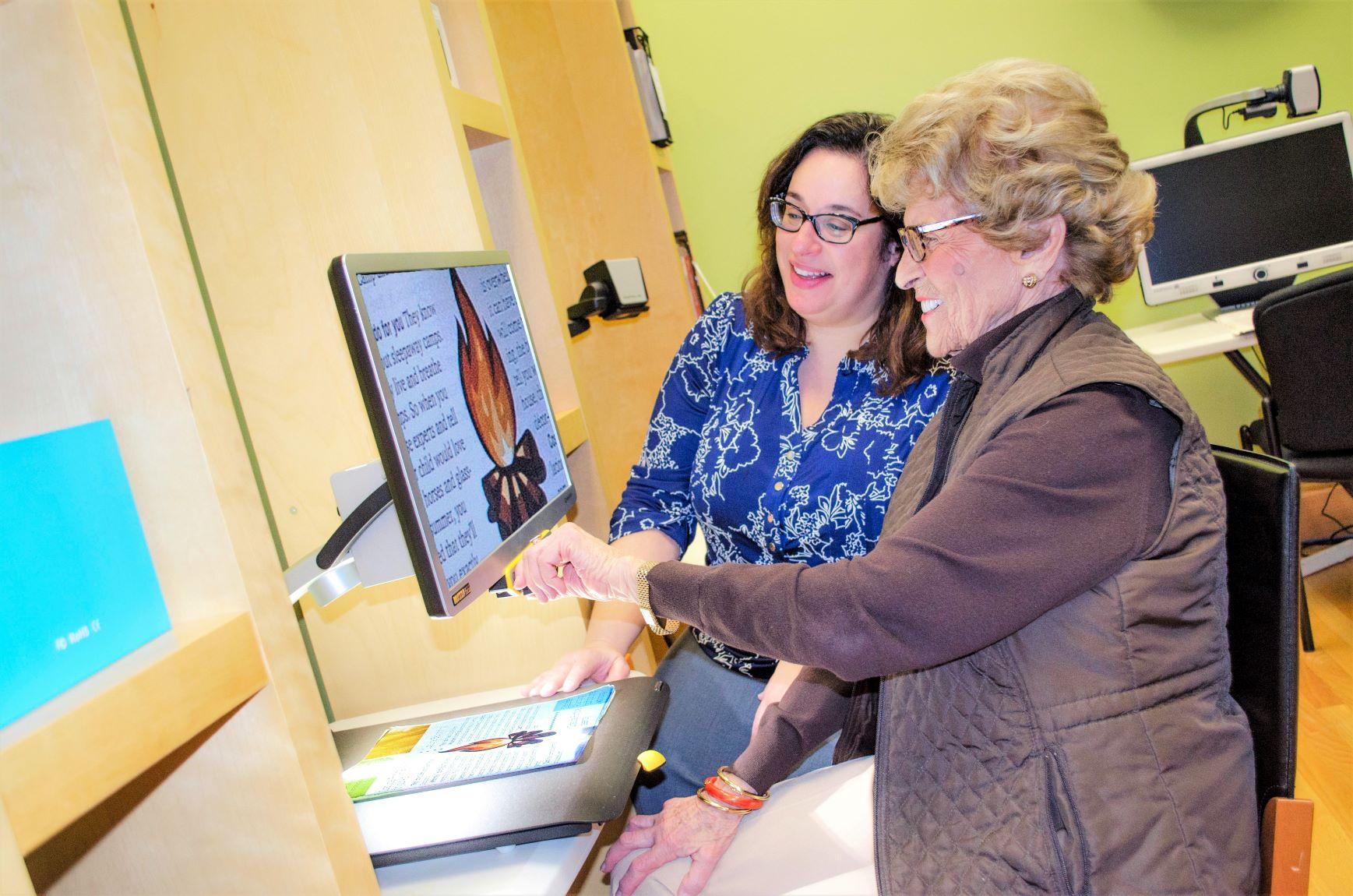 Two women stand together looking at a magnifier which looks like a large  computer screen. An older woman is in the foreground and is reaching out to the screen