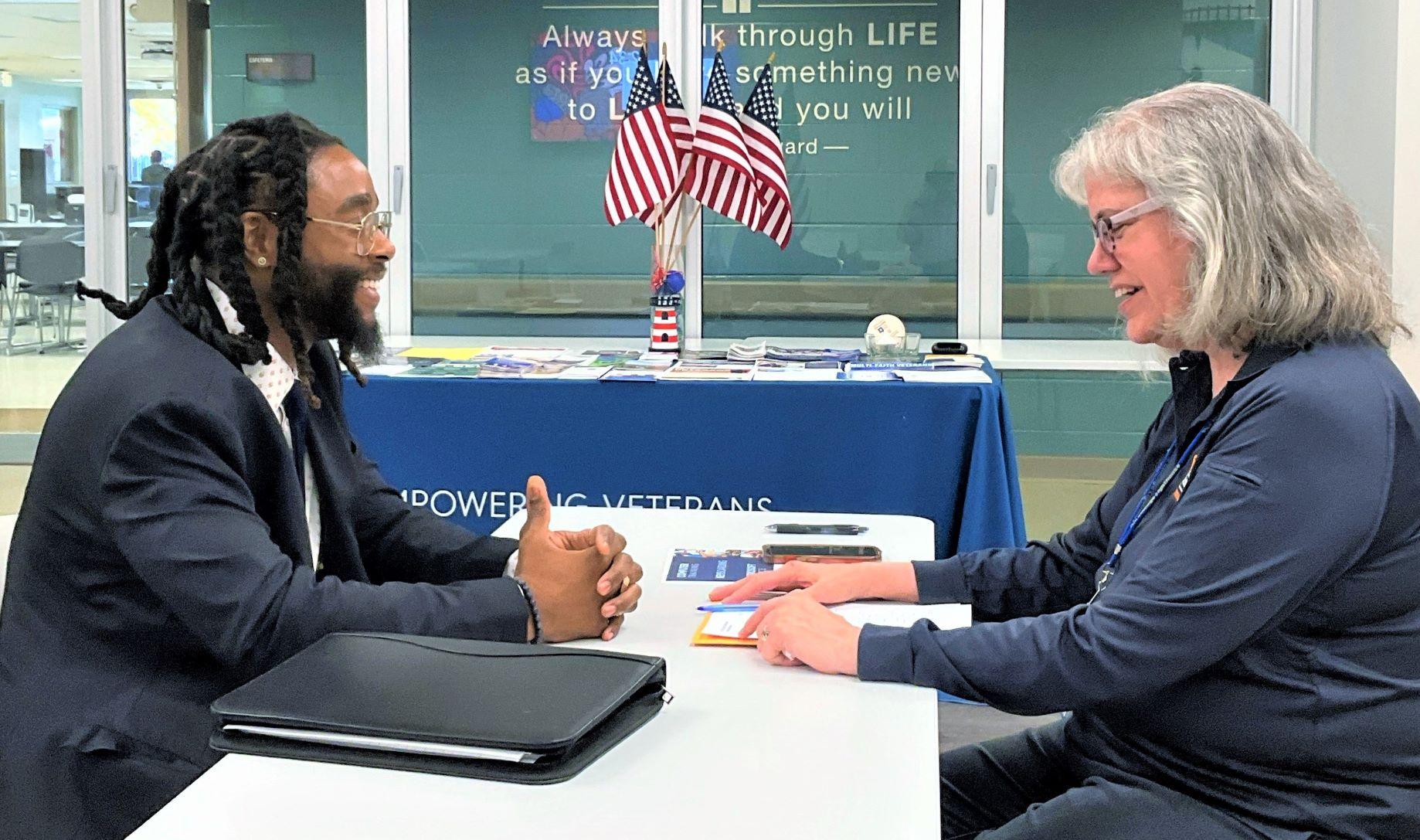 Two individuals are engaged in a practice interview at a table in a brightly lit room with several small American flags in the background.