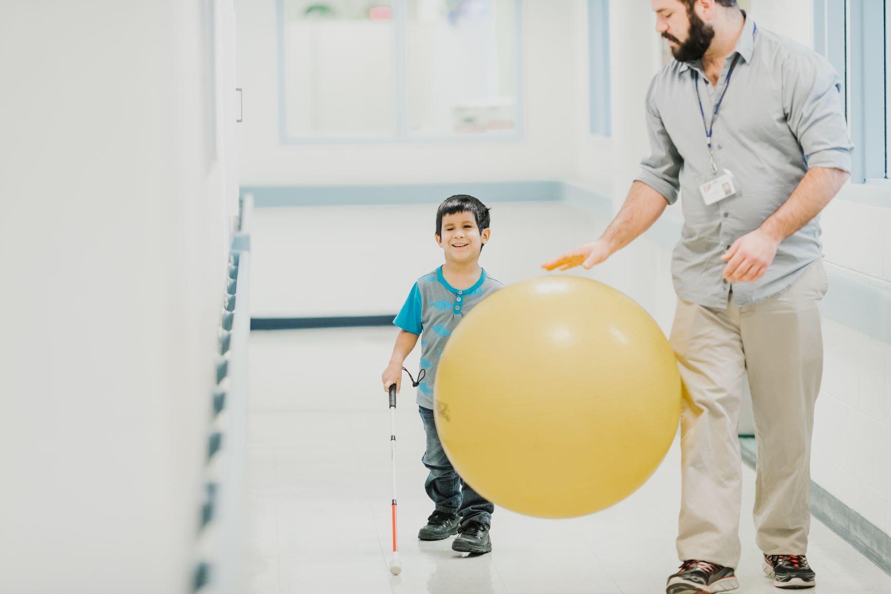 A little boy using a white cane walks down a hallway following a man bouncing a large yellow ball in front of him.